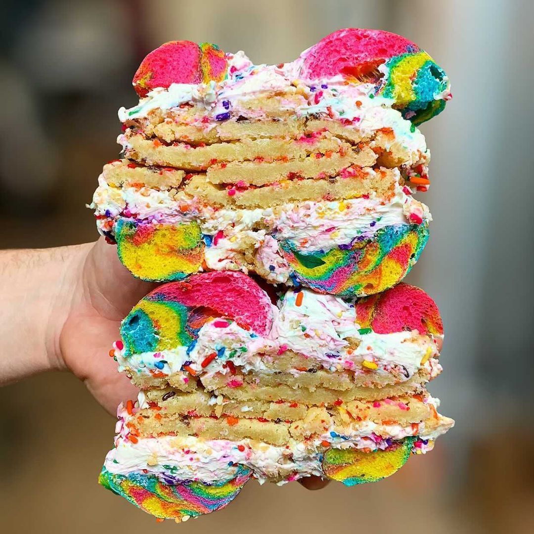 A hand holding stacked rainbow bagel sandwiches loaded with cream cheese and sprinkles